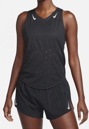 Vrouw draagt een zwart mouwloos geperforeerd sporttopje en zwarte hardloopshorts met Nike-logo's op een witte achtergrond.