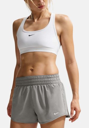 Mujer con un sujetador deportivo blanco de Nike y pantalones cortos deportivos grises de Nike, mostrando el abdomen tonificado y los brazos relajados sobre un fondo liso.