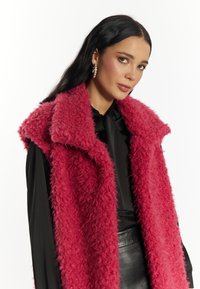 Nepbont vest in felroze met een gestructureerde oppervlakte en een brede schouderkraag, gedragen over een zwarte top en gecombineerd met zwarte leren broek.