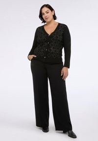 Černý cardigan s diamantovými vzory na přední straně, véčkovým výstřihem a zapínáním na knoflík, doplněný o černé kalhoty širokého střihu a černé boty.