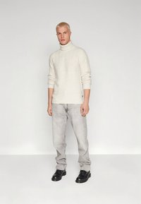 Lichtbeige coltrui met gestructureerd patroon, gecombineerd met lichtgrijze spijkerbroek en zwarte veterlaarzen, staand tegen een neutrale achtergrond.