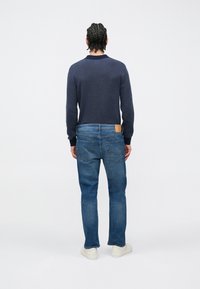 Jersey azul marino con dobladillo y puños acanalados, combinado con jeans de mezclilla azul que cuentan con un parche de cuero y bolsillos estándar, usados con zapatillas blancas.