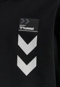 Svart sweatshirt med en tryckt etikett som har texten "Designad av". Nedanför finns två stora chevron-mönster i vitt med prikkig textur.