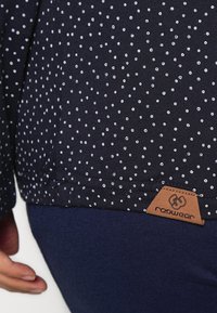 Námorně modrá košile s bílými puntíky, s hnědou koženou dekorací se symbolem 'rapwear' na lemu. Měkká bavlněná tkanina, uvolněný střih.