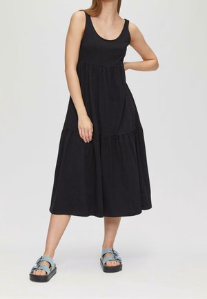 Femme portant une robe midi noire sans manches et des sandales à boucle bleu clair, debout devant un fond uni.