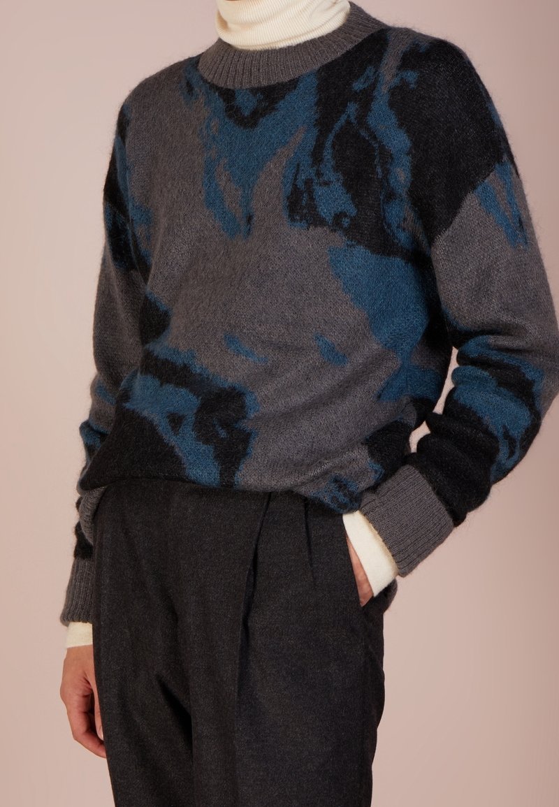 Personne portant un pull à motif abstrait gris et bleu par-dessus un col roulé crème avec un pantalon plissé foncé, main dans la poche.