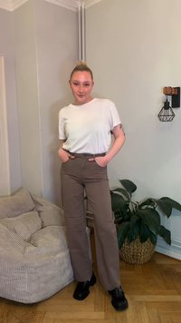 T-shirt branca de manga curta combinada com calças castanhas de cintura alta e sapatos pretos. Ao fundo, um sofá claro e uma planta em vaso.