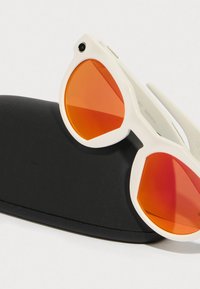 Sonnenbrille mit einem weißen Kunststoffrahmen und orangegetönten Gläsern, die auf einem schwarzen Hartschalenetui liegen. Die Gläser haben eine einzigartige, winklige Form.
