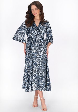 Kobieta ubrana w midi sukienkę w niebiesko-czarny wzór panterki z szerokimi rękawami i paskiem w talii, pozująca z rękami na biodrach na białym tle.