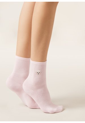 Jambes portant des chaussettes côtelées rose clair avec un petit visage de chien blanc brodé sur le côté, debout sur la pointe des pieds sur un fond uni.