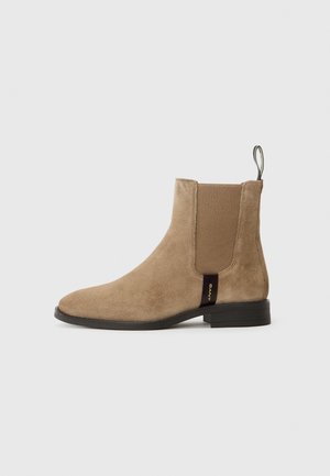 Bottine en suède beige avec des panneaux élastiques sur les côtés et une languette. Semelle plate noire avec une étiquette de marque subtile sur le côté. Silhouette classique.