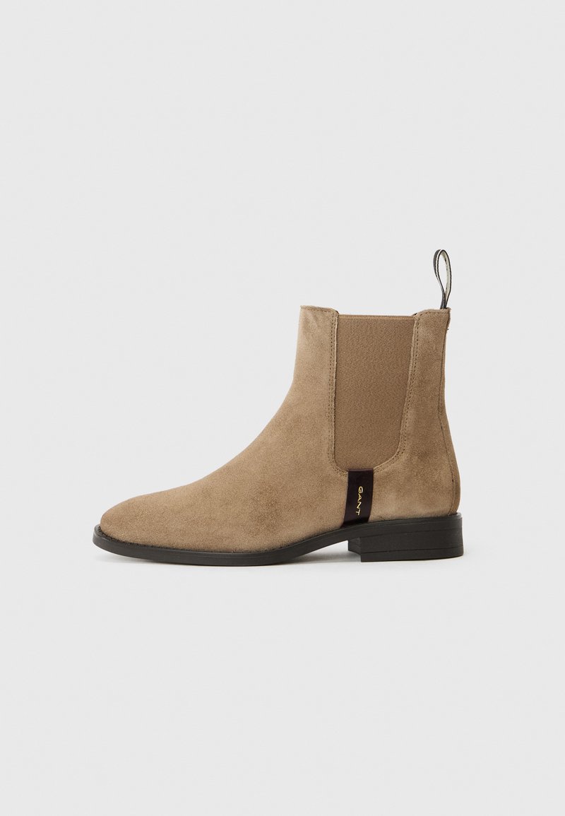 Bottine en suède beige avec des panneaux élastiques sur les côtés et une languette. Semelle plate noire avec une étiquette de marque subtile sur le côté. Silhouette classique.