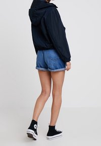 Granatowa, lekka kurtka z kapturem, połączona z podwiniętymi szortami dżinsowymi i czarnymi butami sportowymi za kostkę, widok z tyłu.