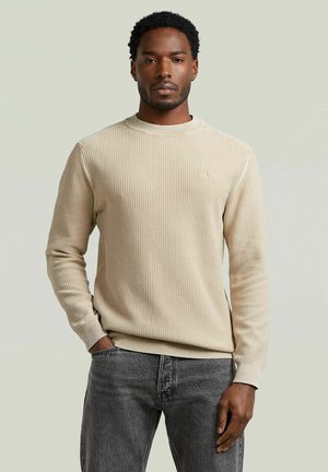 Uomo con capelli corti e ricci che indossa un maglione beige a coste e jeans grigio scuro, in piedi con una mano in tasca davanti a uno sfondo semplice.