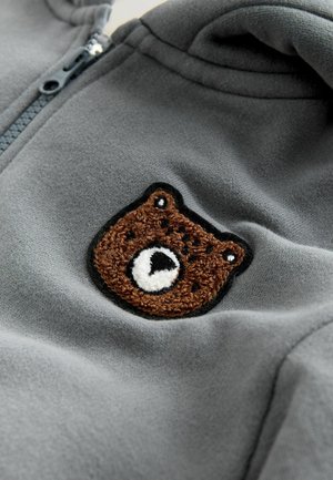 Grå fleece-hoodie med en brunbjörnspatch med svarta konturer, vita accenter och texturerad brodering på bröstområdet.