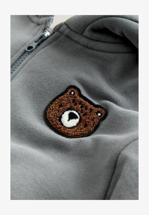 Hallitatud hall fliisist kapuutsiga, mille rinnal on pruun karu paigaldus mustade kontuuride, valgete aktsentide ja tekstuurse tikandiga.