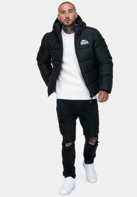 Czarna kurtka puchowa z kapturem, zapinana na zamek, z logo. Nosi ją z białym swetrem z prążkami i zniszczonymi czarnymi dżinsami. Białe sneakersy dopełniają wygląd.