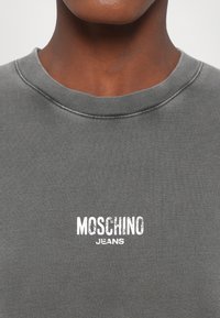 Sudadera gris hecha de tela suave con un cuello acanalado. Tiene un logo blanco prominente que dice "MOSCHINO JEANS" en el pecho.
