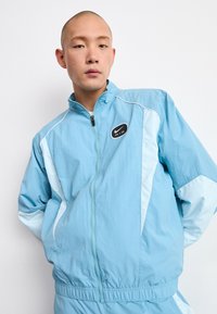 Man i ljusblå Nike Air zip-up jacka med händerna bakom ryggen, poserar mot en plain vit bakgrund.