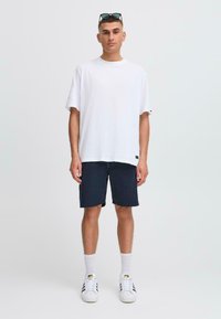 T-shirt en coton blanc, coupe décontractée, associé à un short navy. Le modèle porte des baskets blanches et des chaussettes hautes, des lunettes de soleil reposent sur sa tête.