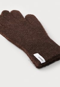 Brown fuzzy knit glove with a white fabric label sewn near the wrist, lying flat on a white surface.