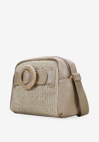 Bolso bandolera beige con frente trenzado, un acento decorativo circular, detalles de tachuelas y cierre de cremallera. Lados de cuero liso.