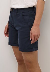 Tmavě modré kraťasy z bavlněné směsi s hladkou texturou, vybavené zapínáním na knoflík, bočními kapsami a ohrnutými lemy.