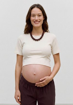 Femme enceinte souriante tenant son ventre nu, portant un haut court clair, un pantalon foncé et un collier épais en perles.