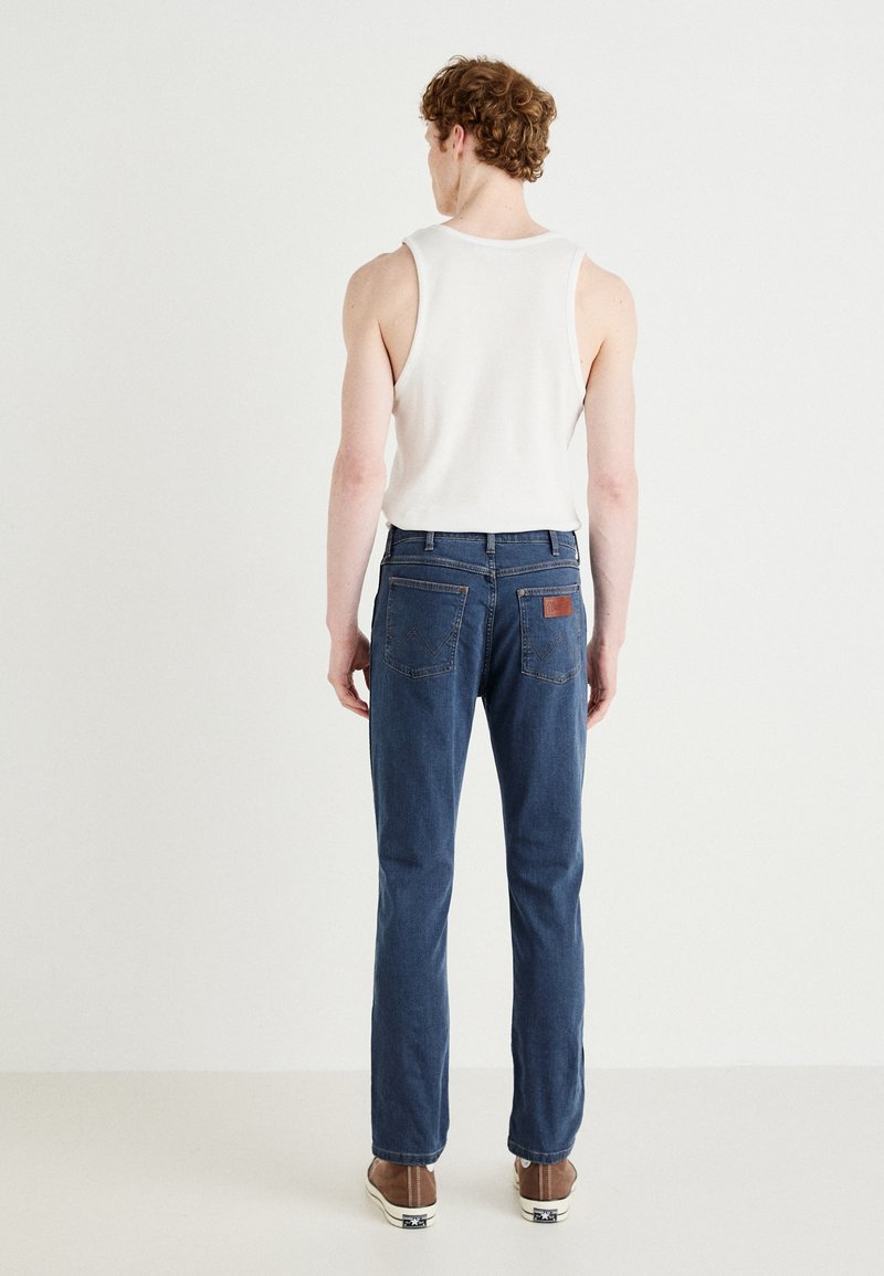 Jeans dżinsowe w ciemnoniebieskim odcieniu, prosty krój z skórzaną metką na tylnej kieszeni, zestawione z białym topem na ramiączkach i brązowym obuwiem.