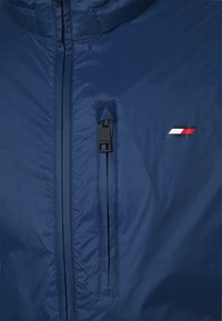 Chaqueta ligera de color azul marino con un bolsillo con cremallera y un acento de logo que presenta rayas rojas, blancas y azules. Material suave y resistente al agua.