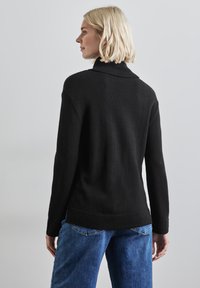 Pull noir avec un col montant, fabriqué dans un tissu doux et texturé, avec un ourlet côtelé. Associé avec un jean en denim bleu.