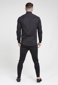 Musta pitkähihainen paita, jossa on napitettava kaulus, istuva malli ja sileä materiaali, yhdistettynä mustiin kapealahkeisiin farkkuihin ja mustiin tennareihin.