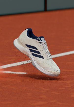 Witte sportschoen met marineblauwe accenten, een mesh-textuur, vetersluiting en zichtbare profielzolen. Midden in de lucht gepositioneerd boven een rode kleibaan.