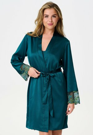 Femme portant un peignoir en satin bleu sarcelle avec des poignets en dentelle florale, noué avec une ceinture assortie, debout devant un fond blanc.