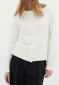 Krémový cardigan vyrobený z měkké pleteniny, s žebrovanou légou na knoflíky, texturálními kontrasty a dlouhými rukávy se žebrovanými manžetami.