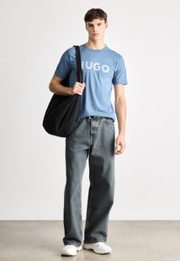 Μπλε βαμβακερό t-shirt με γράμματα "HUGO", χαλαροί ξεθωριασμένοι τζιν, λευκά αθλητικά παπούτσια και μια μαύρη τσάντα ώμου. Απλό φόντο.