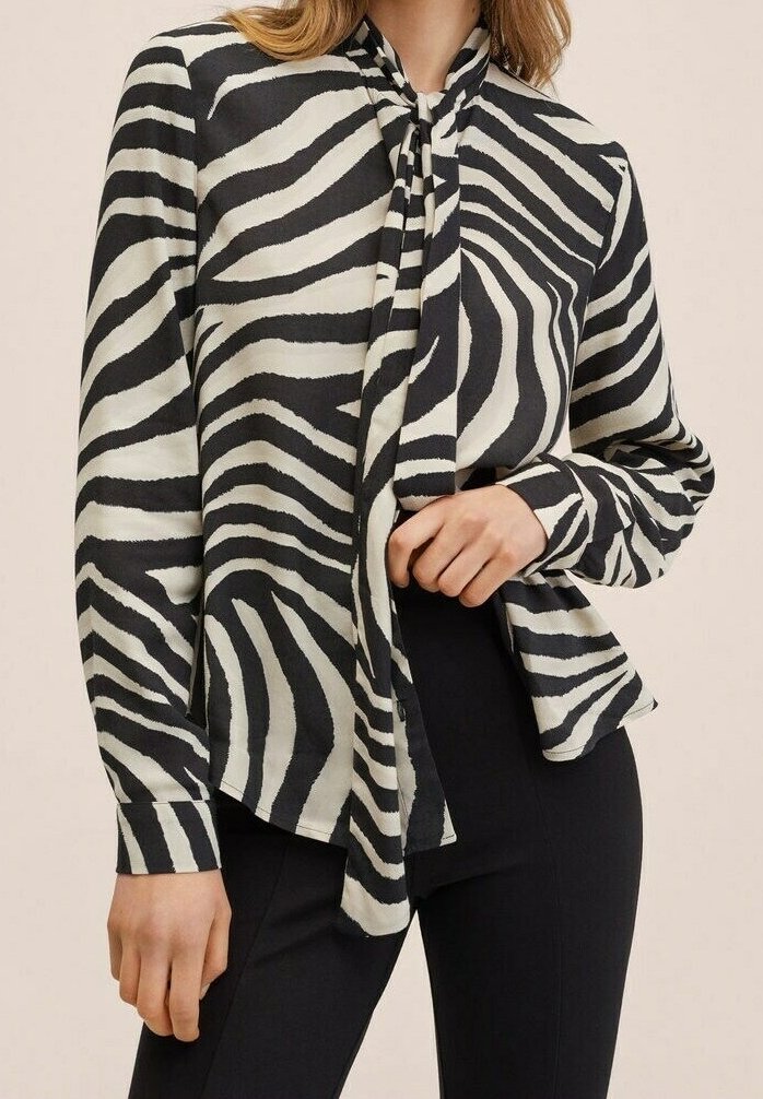 Femme portant une blouse noire et blanche à rayures zébrées avec un col noué en nœud papillon, associée à un pantalon noir ajusté.