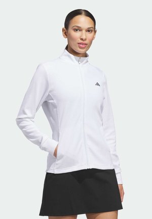 Mujer con una chaqueta blanca con cremallera y un pequeño logo en el pecho y una falda negra, de pie con una mano en el bolsillo.