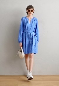 Rochie albastră, cu mâneci lungi, decolteu în v, și talie definită cu o curea, având un model floral texturat. Asortată cu adidași albi și o geantă mică.