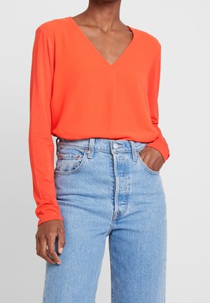 Femme portant un haut orange vif à manches longues avec encolure en V, rentré dans un jean taille haute bleu clair, une main dans la poche.
