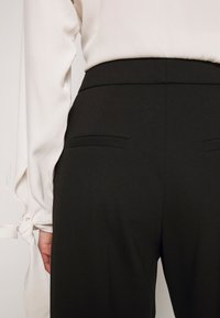 Femme portant un pantalon noir taille haute avec des poches passepoilées à l'arrière et un chemisier blanc à manches longues aux poignets noués.