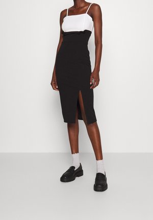 Kobieta ubrana w biały, bezrękawnik oraz czarną ołówkową spódnicę z wysokim stanem i przednim rozcięciem, do tego białe skarpetki i czarne mokasyny.