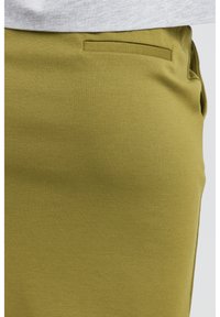Tissu vert olive avec une texture lisse, présentant un devant plat et une seule poche latérale. Gros plan de la zone de taille du vêtement.