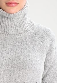 Jasnoszary sweter z golfem o prążkowanej fakturze, z wysokim kołnierzem i rękawami raglanowymi. Materiał wydaje się miękki i potencjalnie ciepły.