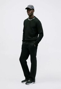 Svart sweatshirt med vit text, svarta byxor och svarta sneakers. Modellen bär en grå keps. Mjuk textur, figurnära design och avslappnad stil.