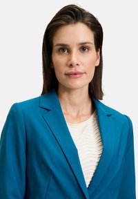 Teal blézer, csúcsos hajtókkal, egy krém színű, textúrázott blúzzal párosítva. A modell közepes barna hajjal rendelkezik, minimális smink van rajta, semleges háttérrel.