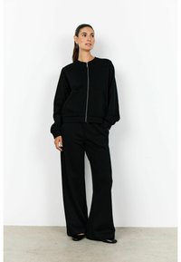 Fekete cipzáras pulóver hosszú ujjakkal és kerek nyakkivágással, széles szárú fekete nadrággal párosítva. Puha anyag, minimalista dizájn.