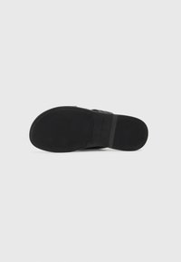 Sandale noire avec une semelle en caoutchouc texturé, dotée de larges lanières et d'un marquage minimal sur le dessous. Le design est plat et ouvert à l'avant.
