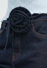 Traper remen s teksturiranim tamnoplavim ukrasom u obliku ruže, s mašnom od vrpce. Vidljivi narančasti šavovi kontrastiraju s tamnim tkanjem.