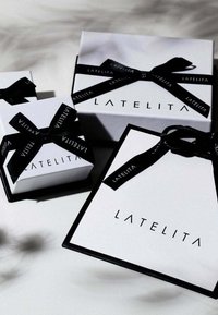 Białe pudełka prezentowe z czarnymi akcentami i wstążkami z napisem "LATELITA". Różne rozmiary ułożone razem, podkreślające elegancki, minimalistyczny design.